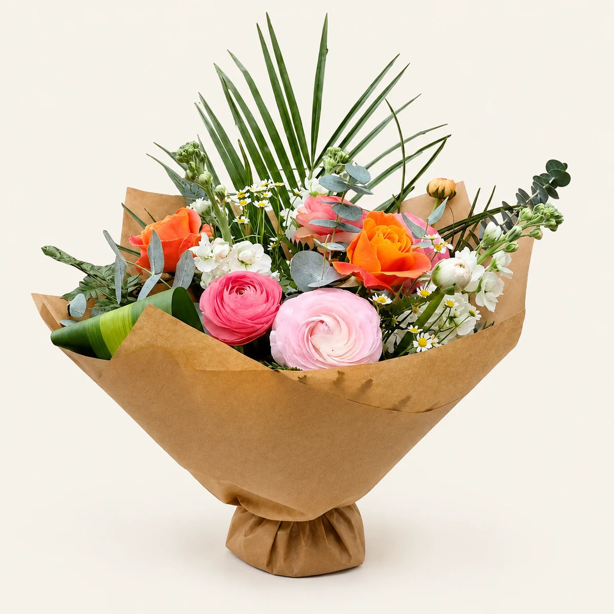 A vibrant floral bouquet wrapped in brown kraft paper, featuring orange and pink roses, pink ranunculus, white daisies, green foliage, and palm leaves, set against a solid off-white background.