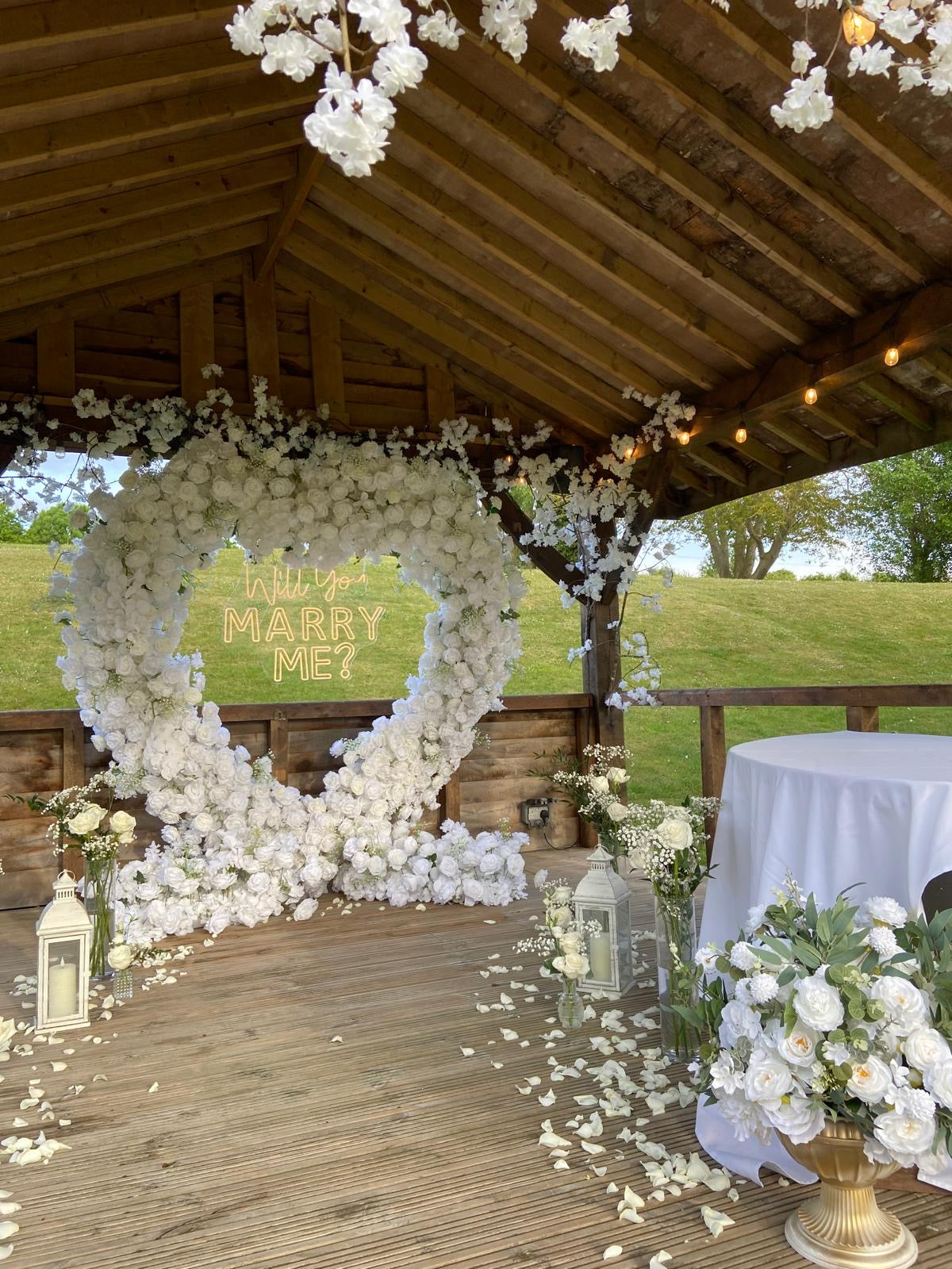 White floral proposal heart with a neon sign, lanterns, candles and floral arrangements in urns.