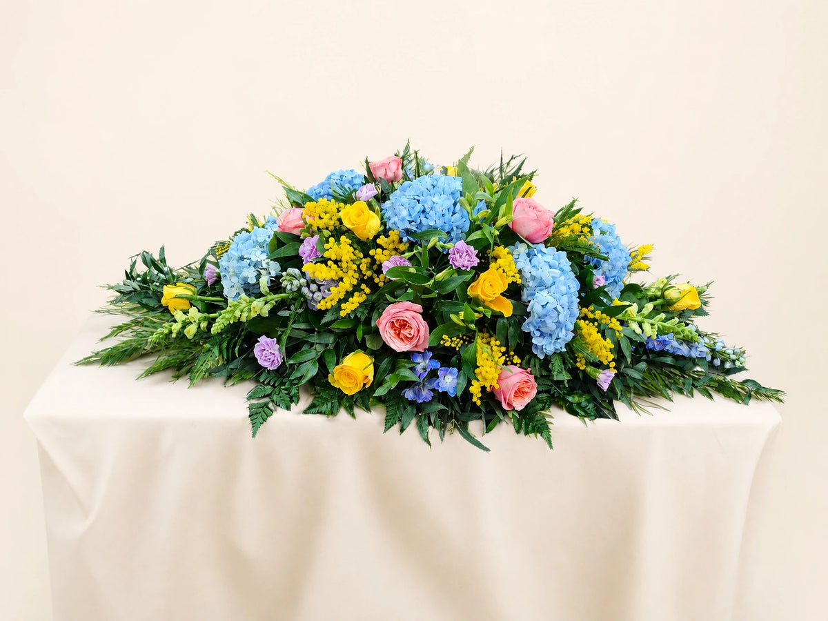 Side view of a funeral casket spray with blue hydrangeas, pink roses, yellow flowers and foliage