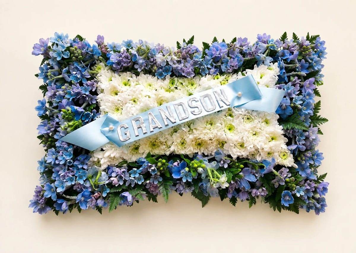 White floral funeral pillow with a blue and purple flower border and a pale blue GRANDSON ribbon