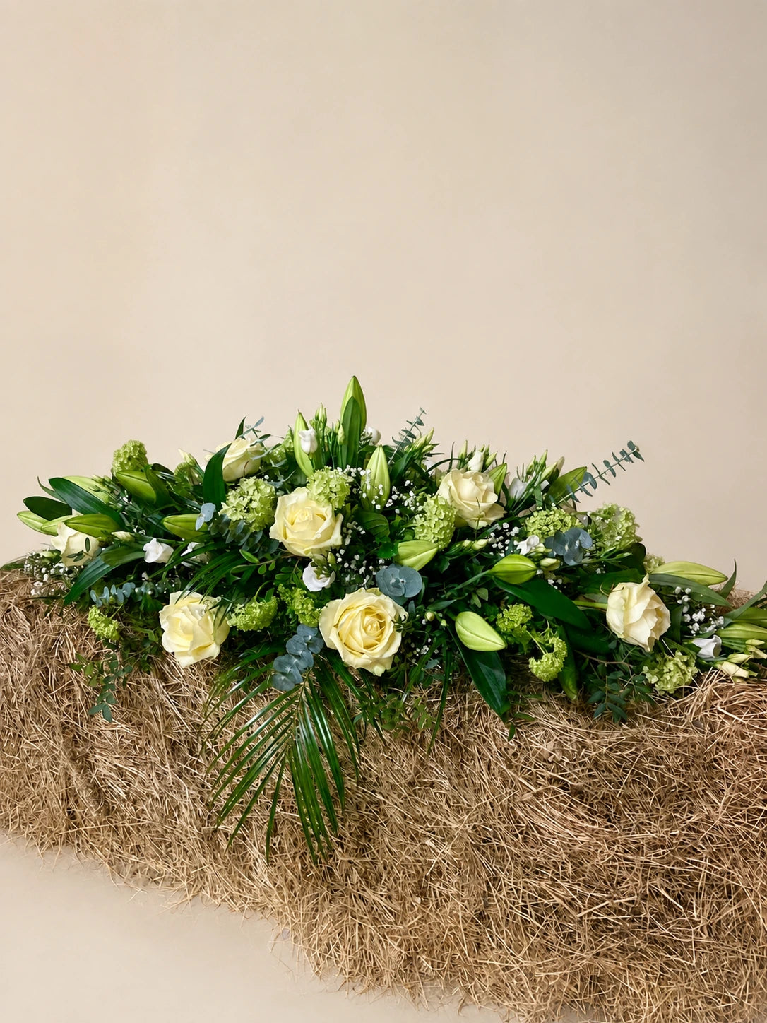 Side view of a cream rose and lily casket spray displayed on a straw bale