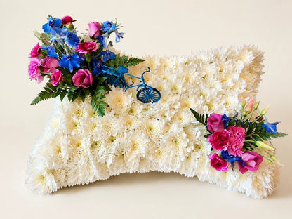 White floral funeral pillow with pink roses, blue flowers and foliage at opposite corners