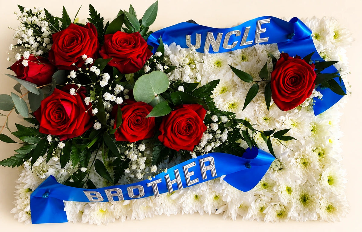 White floral funeral pillow with red roses and blue ribbons reading UNCLE and BROTHER