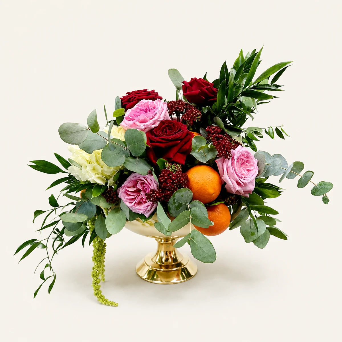 A floral arrangement in a gold pedestal bowl, featuring red and pink roses, green eucalyptus leaves, white flowers, and two oranges, photographed on a clean off-white background.