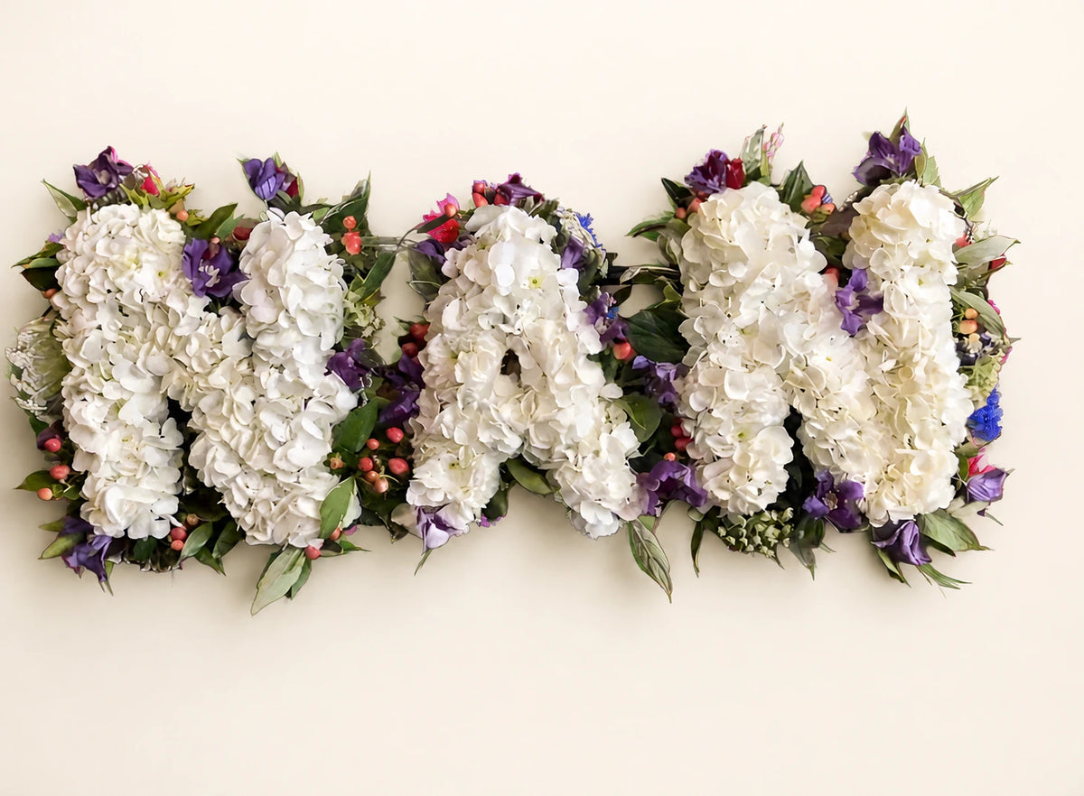 White hydrangea funeral letters spelling NAN surrounded by purple flowers and greenery