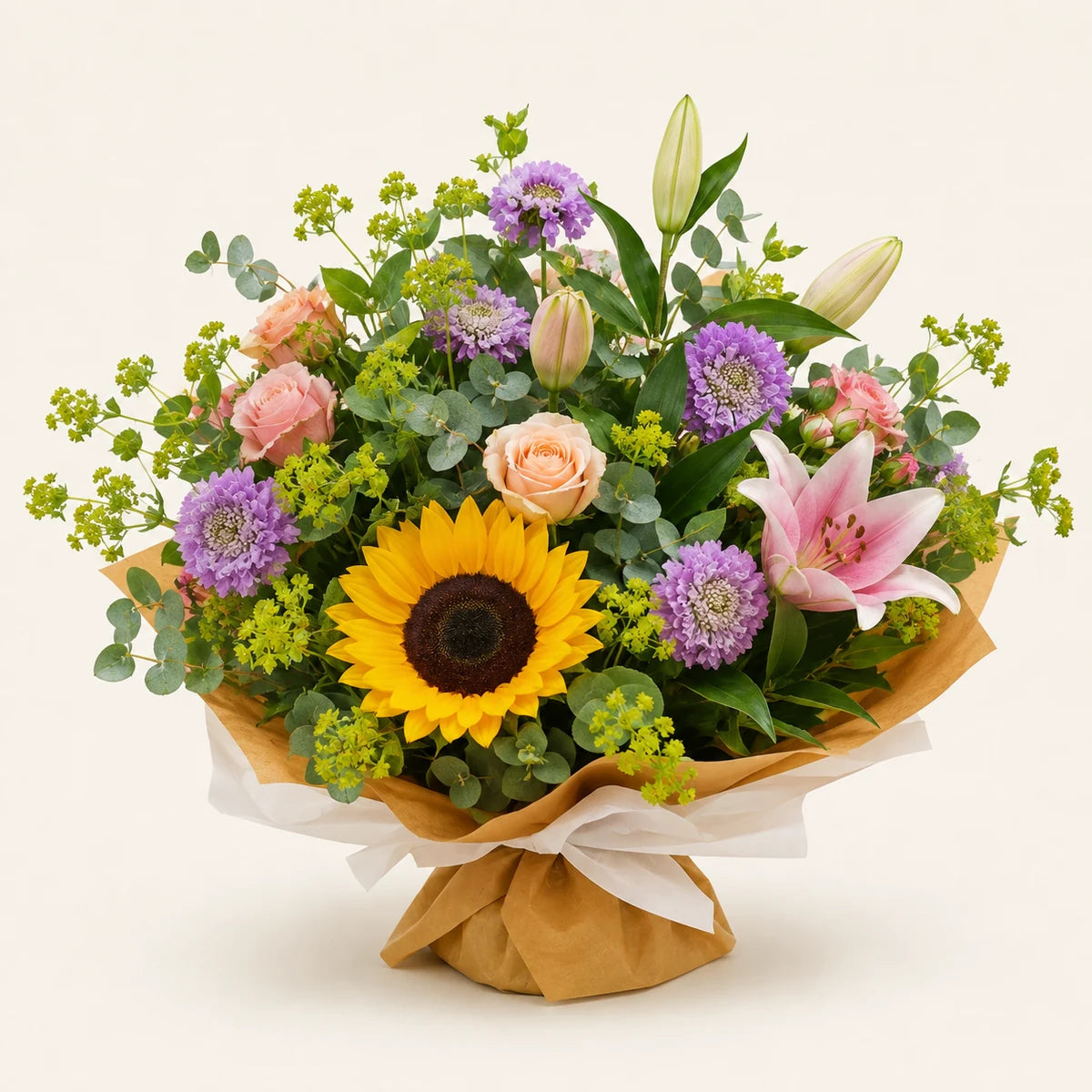 A vibrant mixed floral bouquet wrapped in brown kraft paper and white ribbon, featuring a large yellow sunflower, pink lily, purple flowers, peach roses, and green foliage on a light cream background.