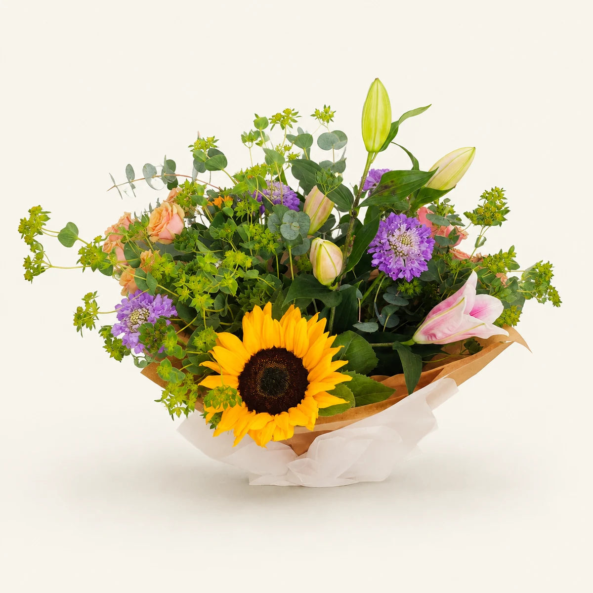 A mixed floral bouquet featuring a large yellow sunflower, purple flowers, pink lilies, green foliage, and pale yellow buds, wrapped in brown paper and white tissue paper against a solid off-white background.