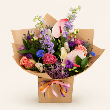 A vibrant mixed flower bouquet wrapped in brown kraft paper and tied with a pink, orange, and purple ribbon, set against a solid light beige background.