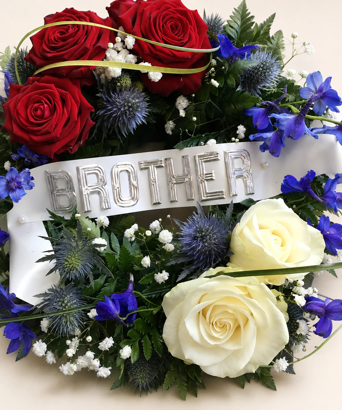 Close view of a funeral wreath with red and cream roses, blue thistles and a ribbon reading BROTHER