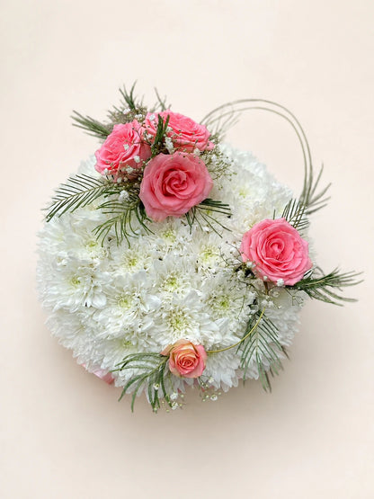 Round white floral funeral posy with pink roses, green foliage and curved decorative grasses