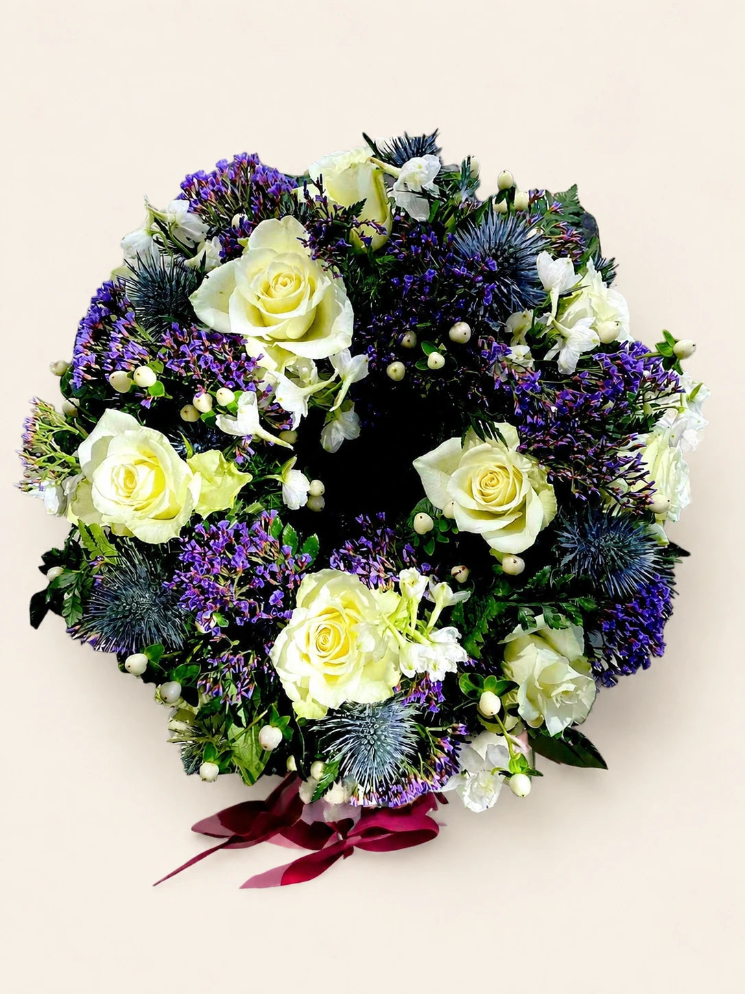Funeral wreath with cream roses, blue thistles, purple flowers and a dark red ribbon