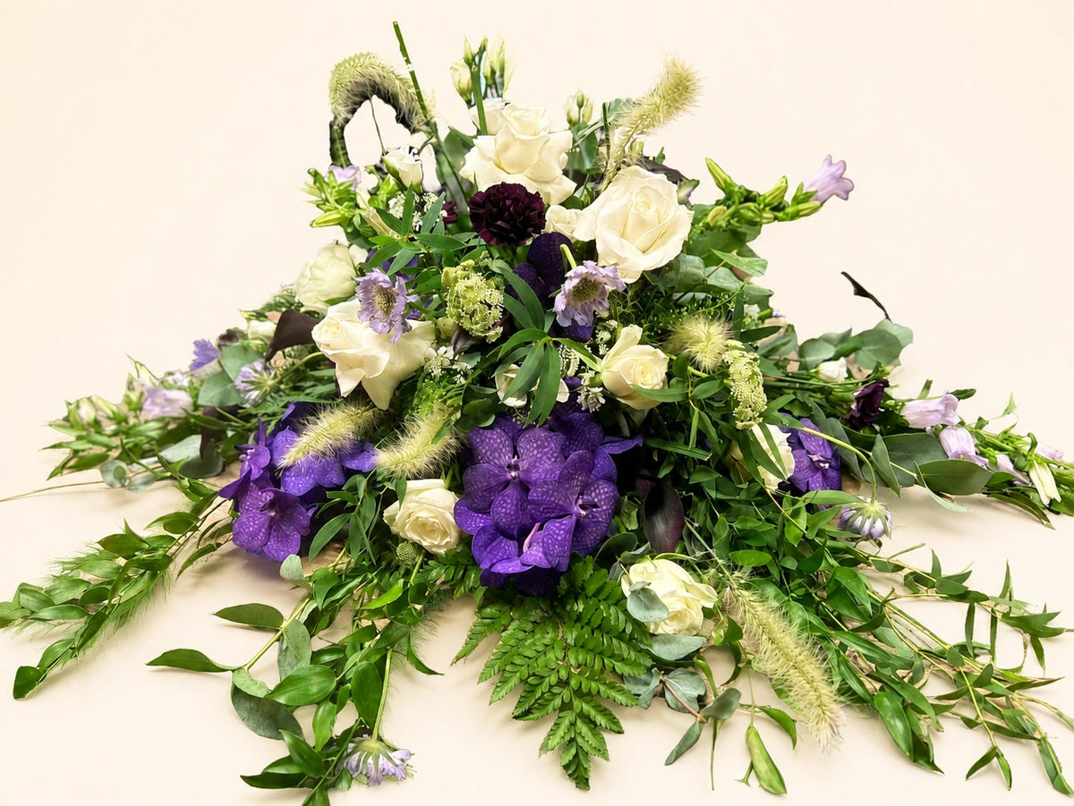 Side view of a funeral posy with purple flowers, cream roses and loose trailing green foliage