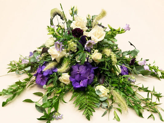 Side view of a funeral posy with purple flowers, cream roses and loose trailing green foliage