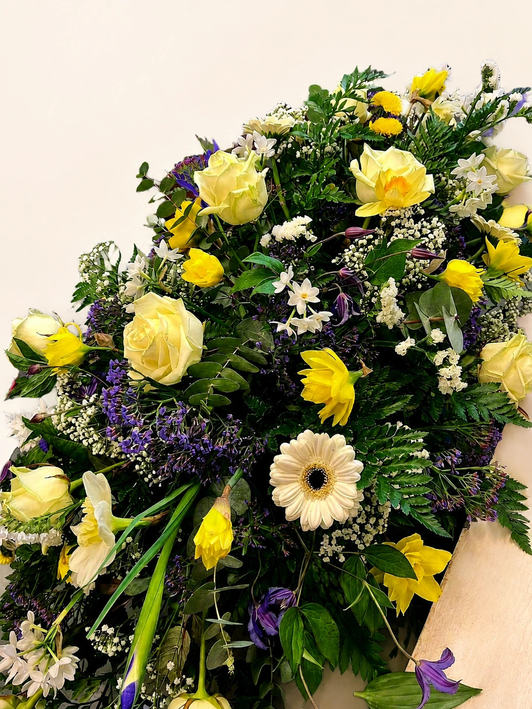 Angled view of a funeral casket spray with yellow roses, white gerberas and purple flowers