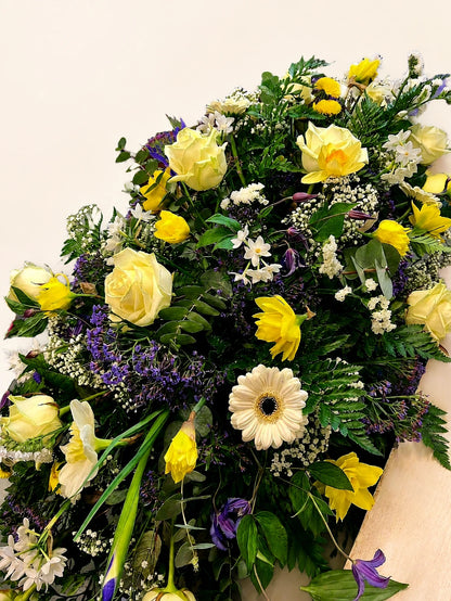 Angled view of a funeral casket spray with yellow roses, white gerberas and purple flowers