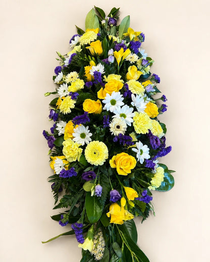 Overhead view of a yellow, white and purple funeral casket spray with green foliage