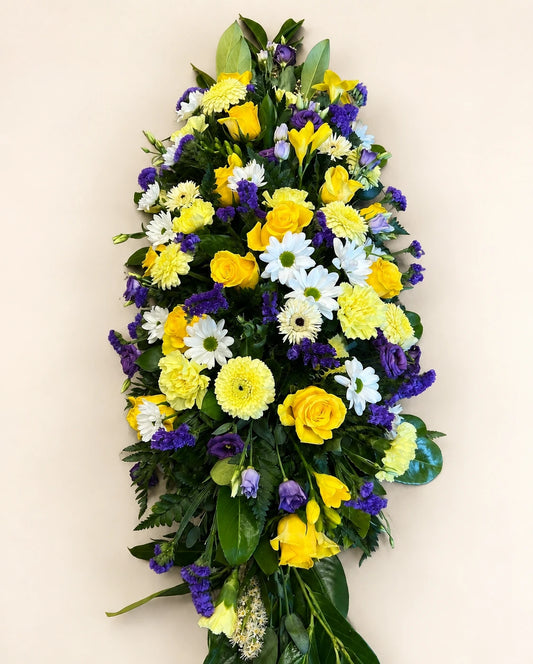 Overhead view of a yellow, white and purple funeral casket spray with green foliage