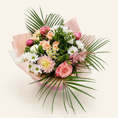 A vibrant floral bouquet featuring pink and white flowers, green foliage, and long palm leaves, wrapped in light pink tissue paper against a solid off-white background.