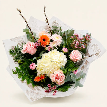 A wrapped floral bouquet featuring pink roses, a large white hydrangea, orange gerbera daisies, pink daisies, green foliage, and small pink berries, arranged in white patterned paper on a light cream background.