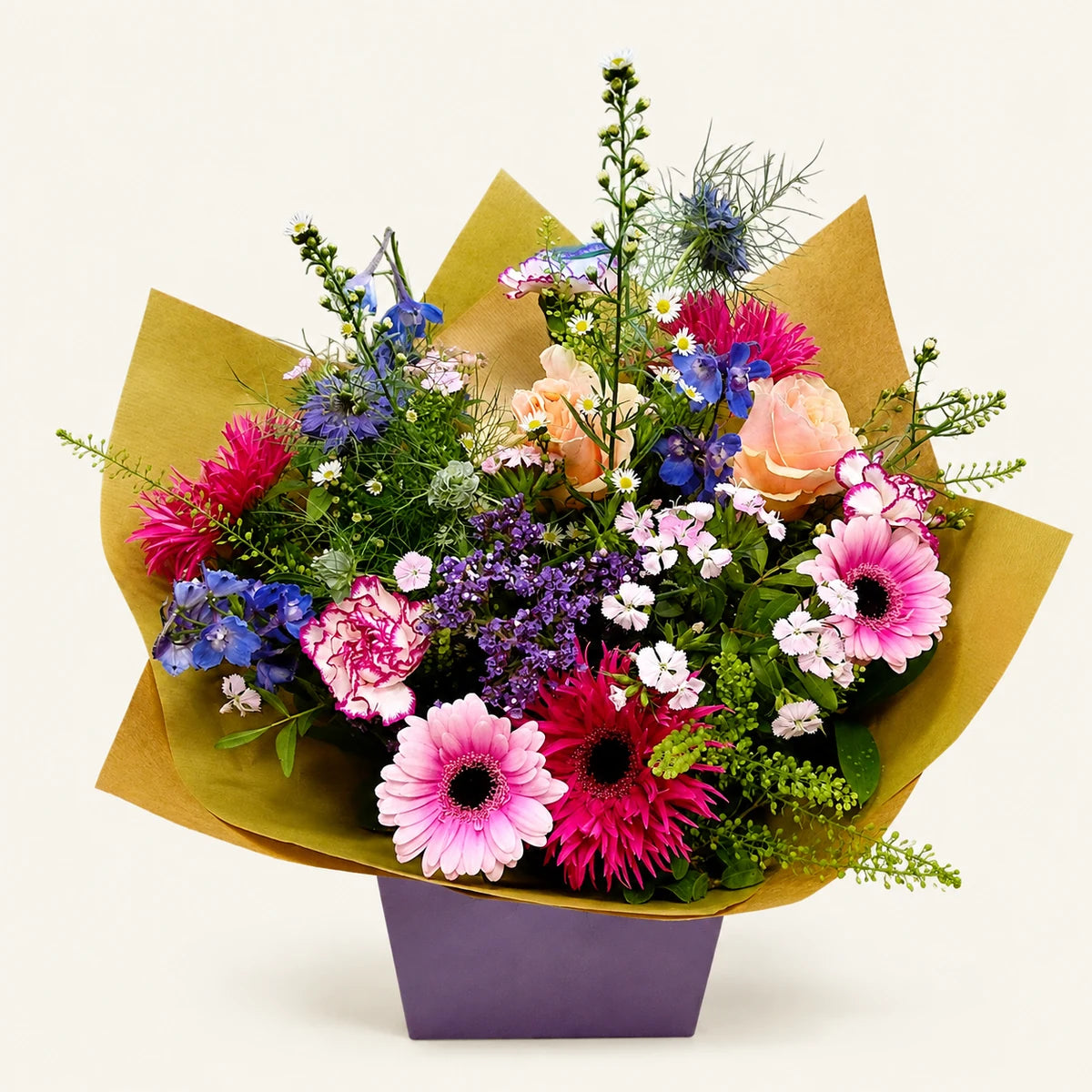 A vibrant mixed flower bouquet wrapped in gold paper and presented in a purple box, photographed against a clean, light off-white background.
