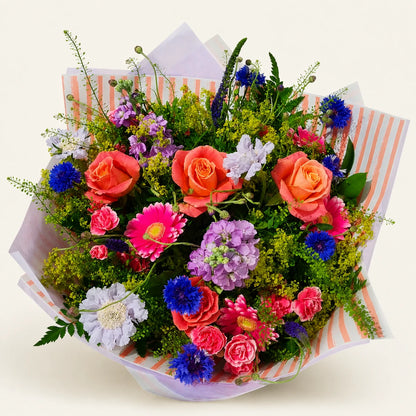 A vibrant, multicolored floral bouquet featuring orange roses, pink gerbera daisies, purple and blue flowers, and green foliage, wrapped in striped paper on a light cream background.