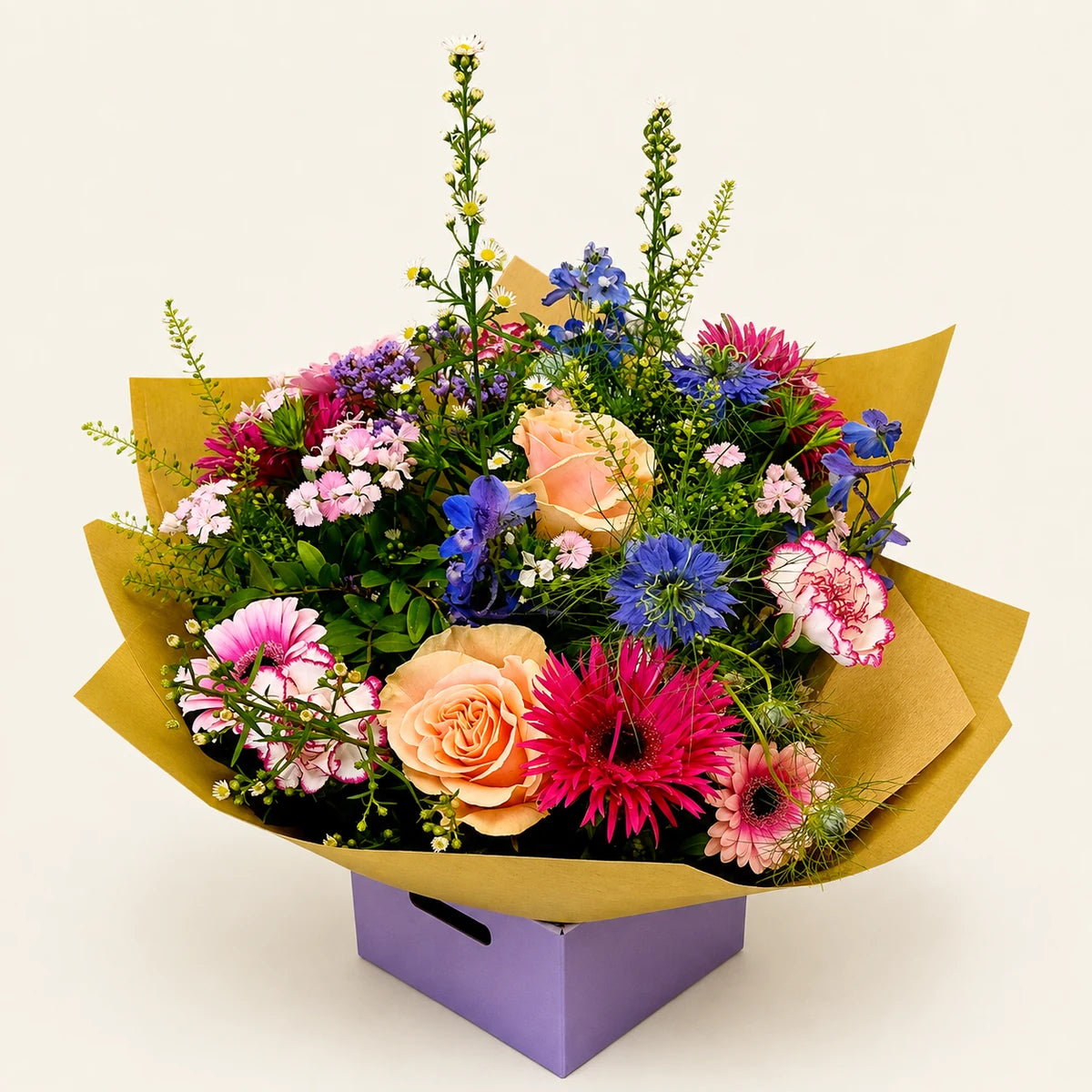 A vibrant mixed flower bouquet wrapped in gold paper and presented in a purple box, featuring pink, orange, blue, and purple flowers on a light cream background.