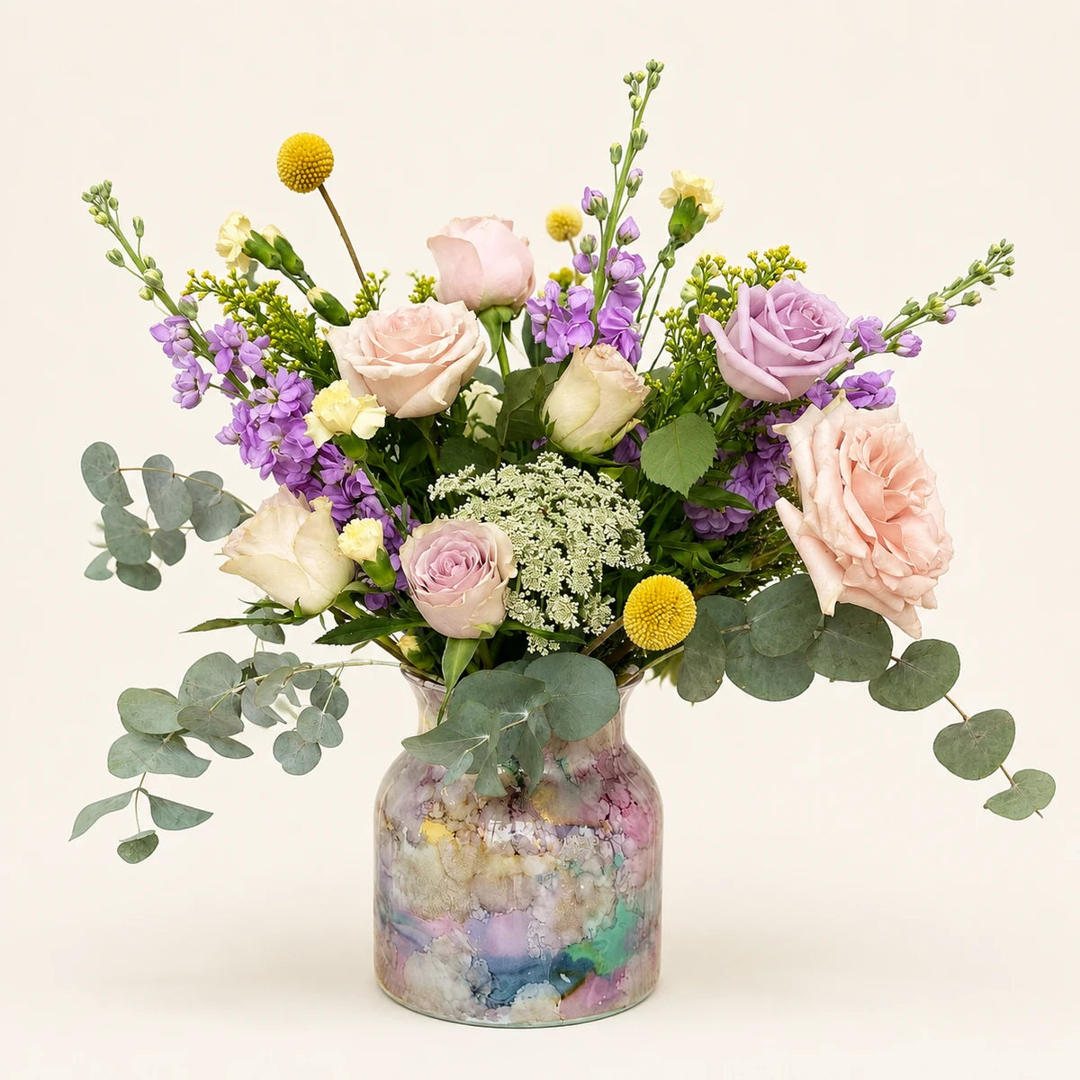 A vibrant floral arrangement featuring light pink and lavender roses, purple stock, yellow globe flowers, and green eucalyptus leaves, presented in a clear glass jar filled with pastel-colored pebbles on a warm off-white background.