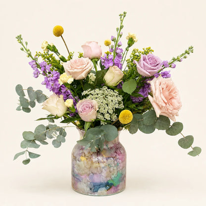 A vibrant floral arrangement featuring light pink and lavender roses, purple stock, yellow globe flowers, and green eucalyptus leaves, presented in a clear glass jar filled with pastel-colored pebbles on a warm off-white background.