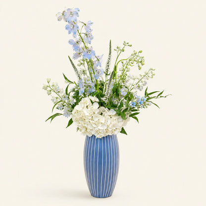 A floral arrangement featuring white hydrangeas, light blue delphiniums, and green foliage in a blue ribbed ceramic vase, set against a solid off-white background.