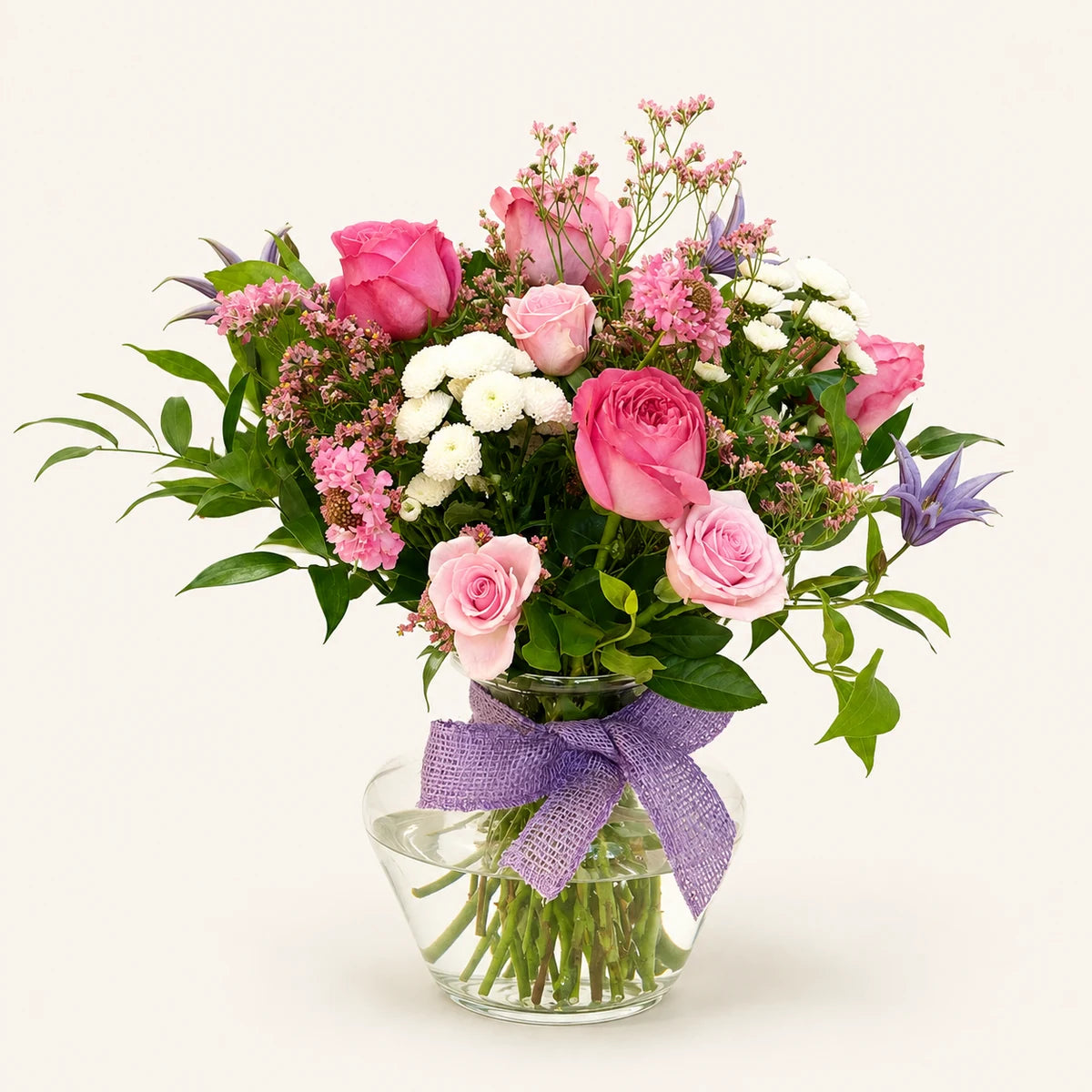 A vibrant floral bouquet featuring pink and light pink roses, white baby's breath, green foliage, and a purple ribbon tied around a clear glass vase, set against a solid off-white background.