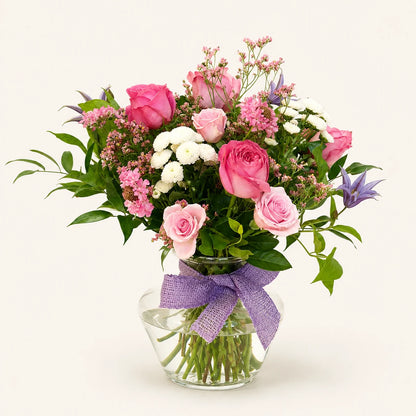 A vibrant floral bouquet featuring pink and light pink roses, white baby's breath, green foliage, and a purple ribbon tied around a clear glass vase, set against a solid off-white background.