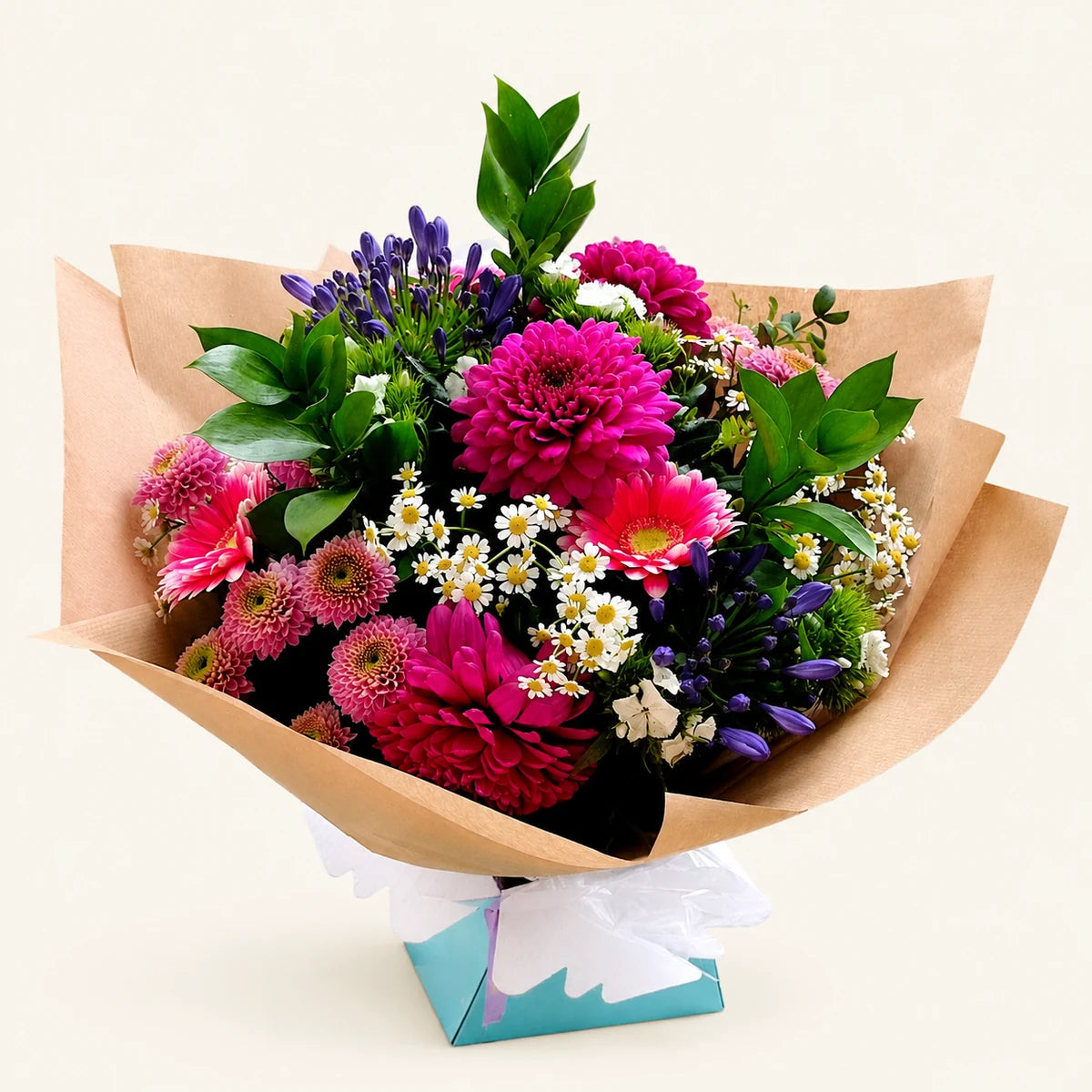 A vibrant mixed flower bouquet wrapped in brown kraft paper and a small teal gift box with white ribbon, featuring bright pink and magenta flowers, green foliage, and small white daisies on a light beige background.
