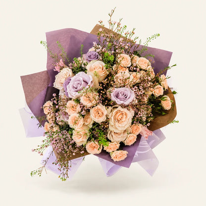 A beautifully arranged floral bouquet featuring light purple and peach roses, mixed with delicate baby's breath and green foliage, wrapped in brown paper and sheer lavender ribbon against a solid off-white background.