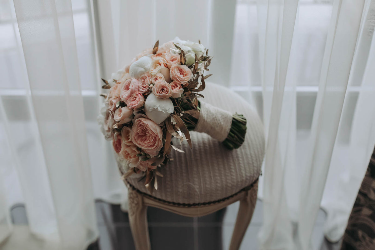 Blush rose bridal bouquet with a ribbon-wrapped handle resting on a white woven chair.