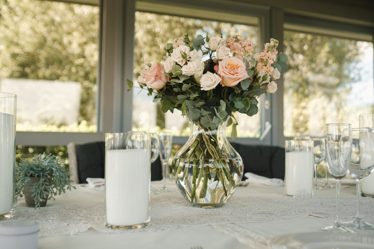 Blush roses and white flowers in a clear glass vase, surrounded by candles on a wedding table.