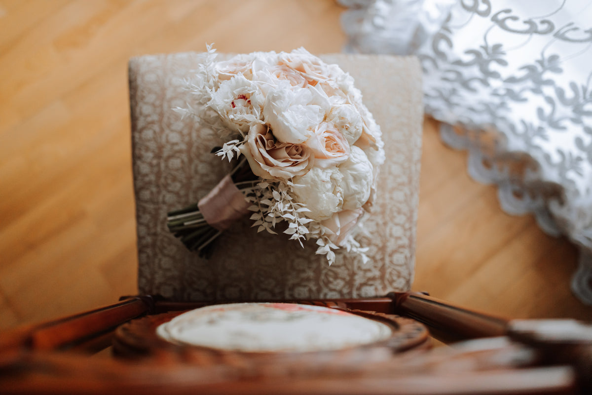 Cream and blush wedding bouquet resting on a patterned chair beside a window.