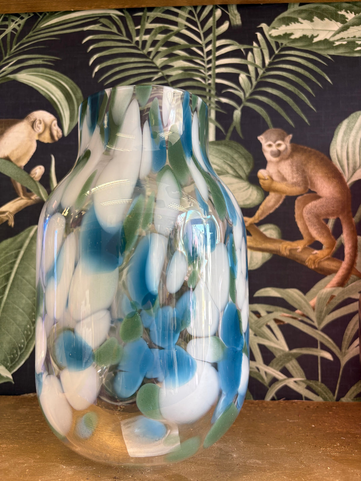 Clear glass vase with blue, green and white oval patterns, displayed against botanical wallpaper