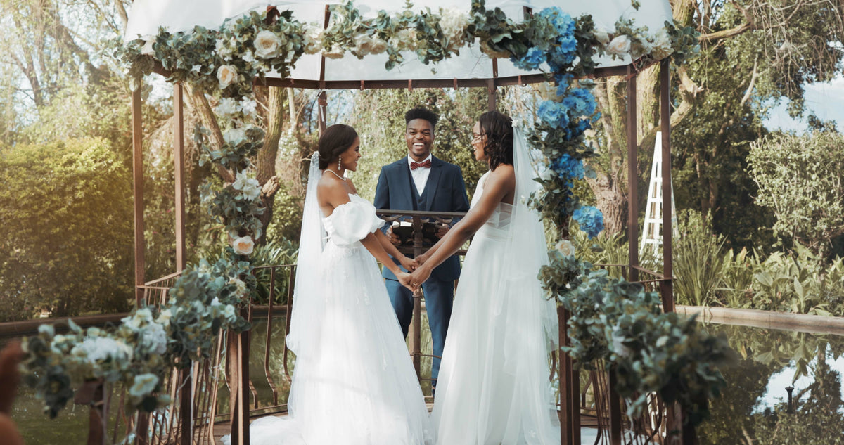 Two brides holding hands during an outdoor ceremony beneath a floral arch, with their celebrant.