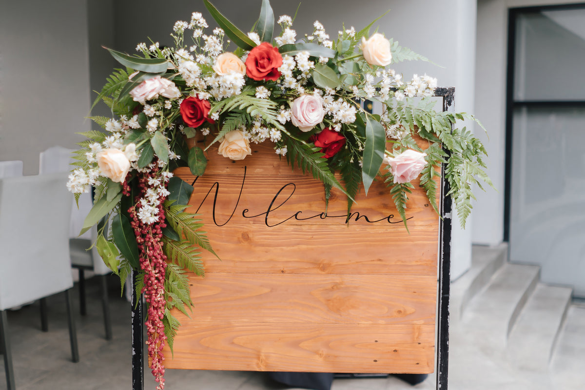 Wooden wedding welcome sign framed by peach, pink and red flowers with trailing foliage.