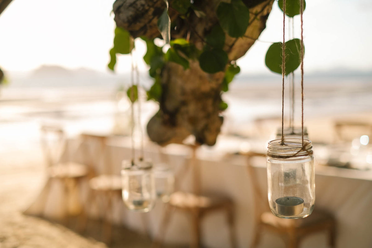 Clear glass candle jars hanging from a tree decorated with green leaves at an outdoor wedding.