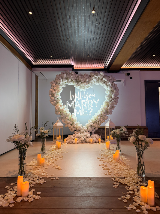 White floral heart and Will you marry me sign with lanterns, candles and petals in an indoor proposal setup.