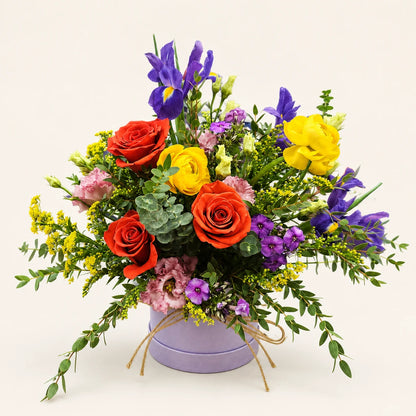 Orange roses, purple irises and yellow ranunculus with pink flowers and eucalyptus in a lilac Signature Hatbox