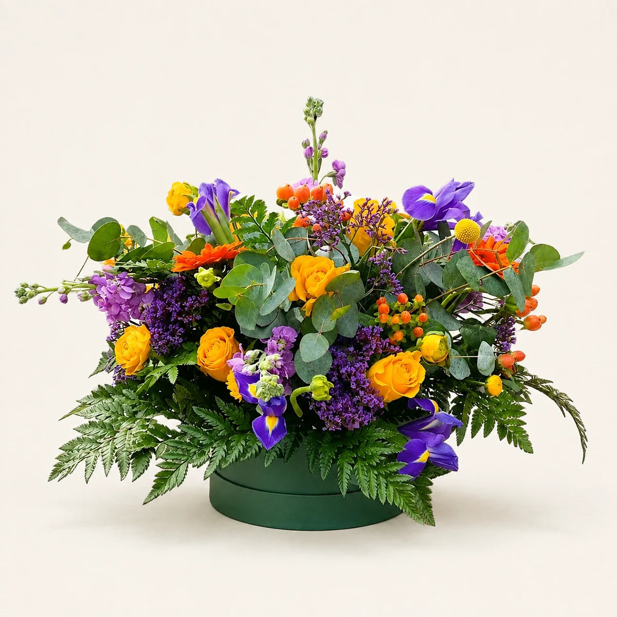 Yellow roses, purple irises and stock with orange berries and eucalyptus arranged in a dark green Signature Hatbox