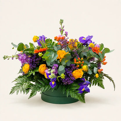 Yellow roses, purple irises and stock with orange berries and eucalyptus arranged in a dark green Signature Hatbox