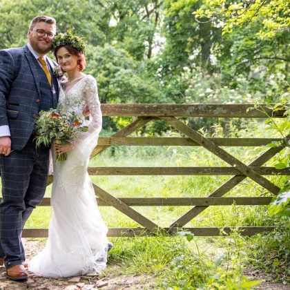 Bride and groom beside a wooden gate in woodland, with a natural green and white bridal bouquet.