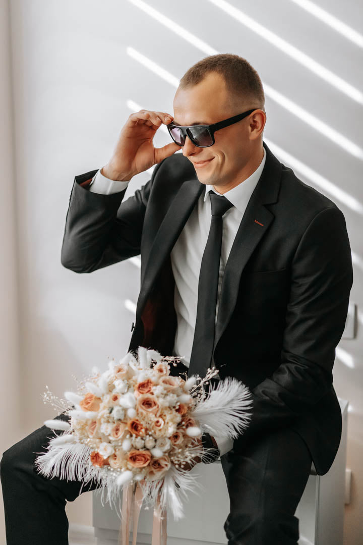 Groom in a dark suit and sunglasses holding a pale peach rose and pampas wedding bouquet.