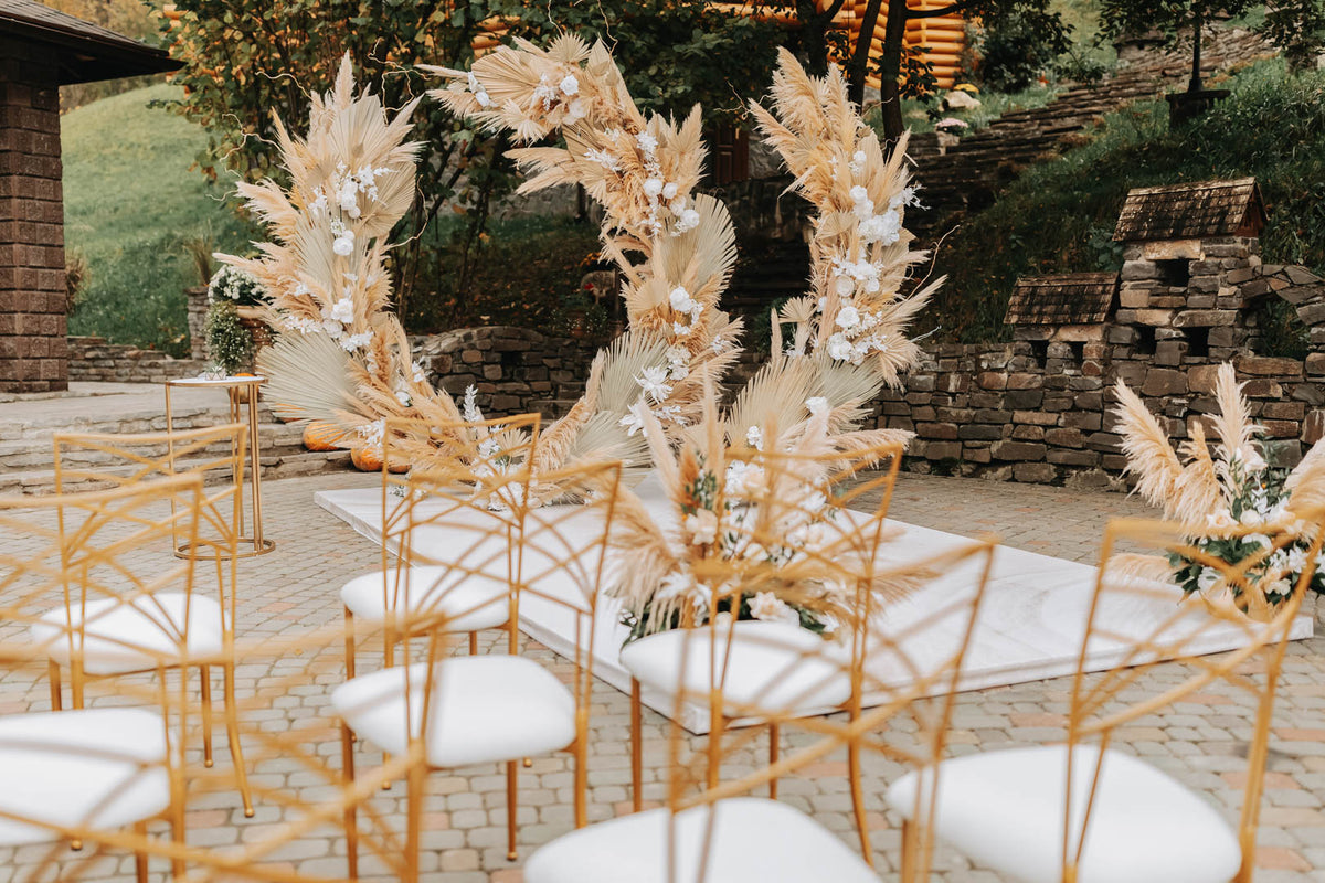 Large circular pampas and dried flower arch beside gold chairs at an outdoor wedding ceremony.