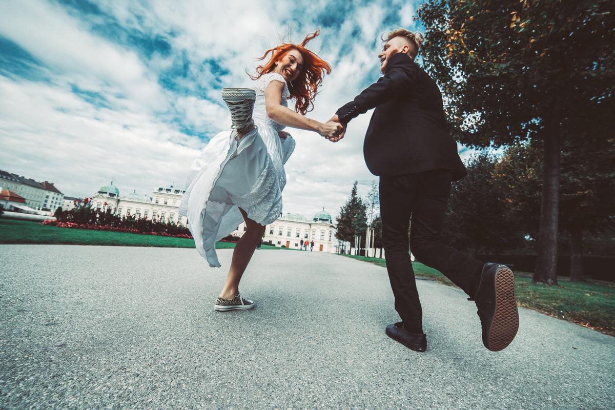 Bride and groom holding hands and jumping outside a wedding venue.