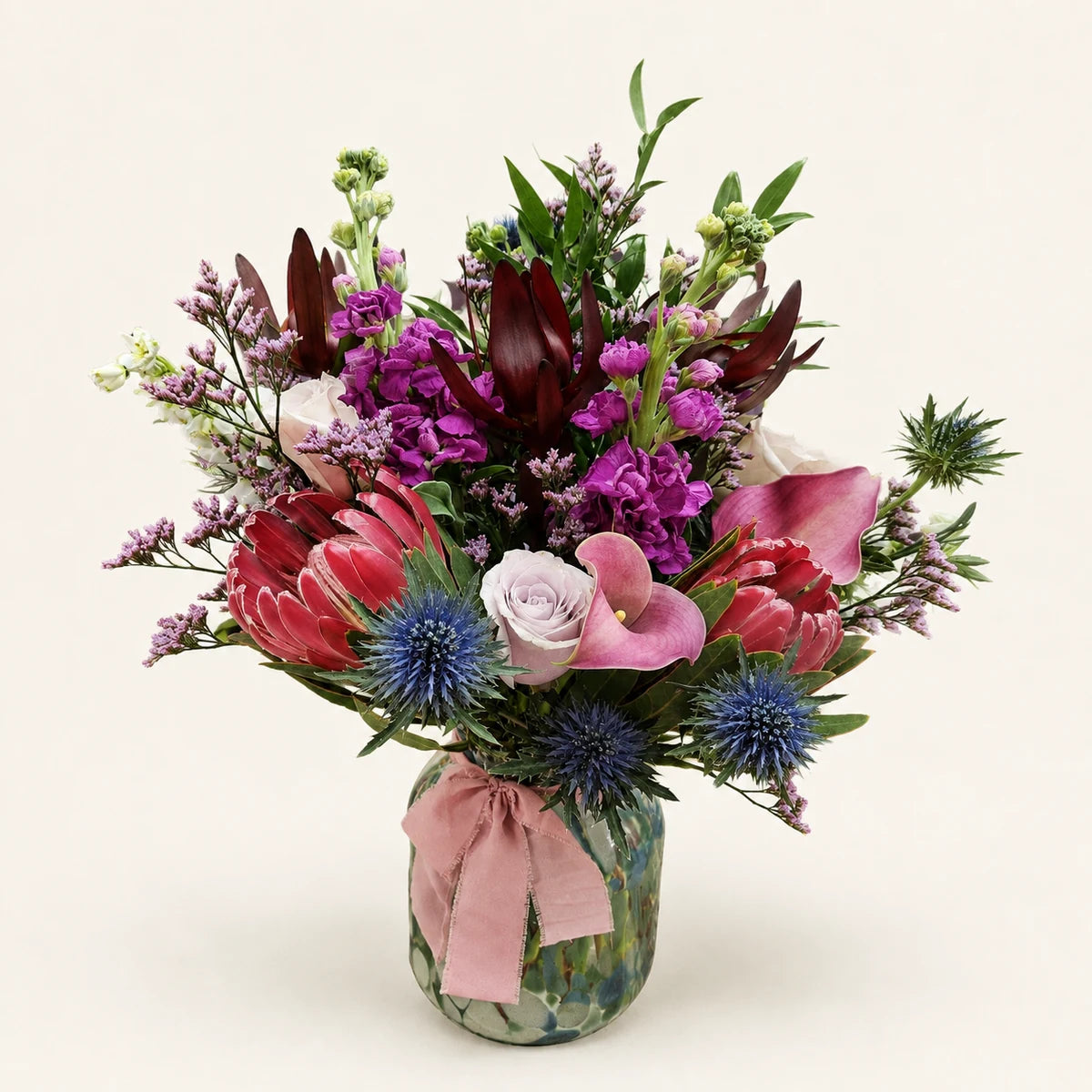 Pink proteas, calla lilies, purple stock and blue thistles in a patterned vase tied with a pink ribbon