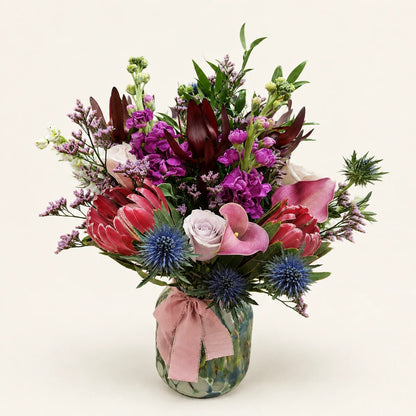 Pink proteas, calla lilies, purple stock and blue thistles in a patterned vase tied with a pink ribbon